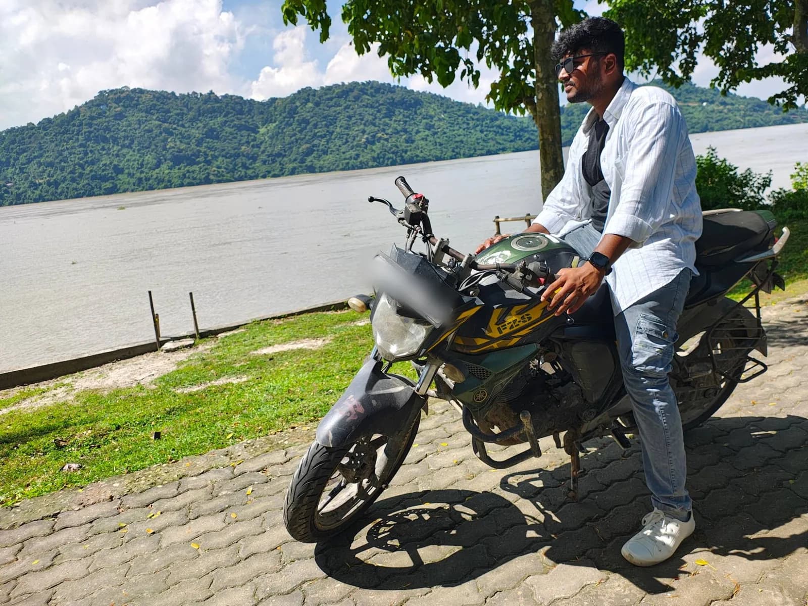 A rider on our Yamaha FZ-S rental bike beside a wide river, with forested hills behind